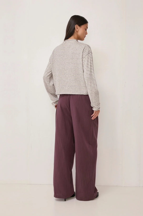 A woman is shown from the back wearing a gray and white horizontally striped long-sleeve crop top and wide-leg mauve purple trousers, standing against a plain white wall.