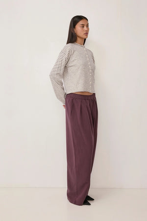 A woman stands in profile wearing a cropped, long-sleeve striped knit top in beige and white, paired with loose, wide-leg mauve-purple trousers, against a plain white studio background.