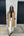 A woman stands outdoors in front of a concrete wall and black garage doors, wearing a long tan trench coat over a black top and white wide-leg pants with black pointed flats.
