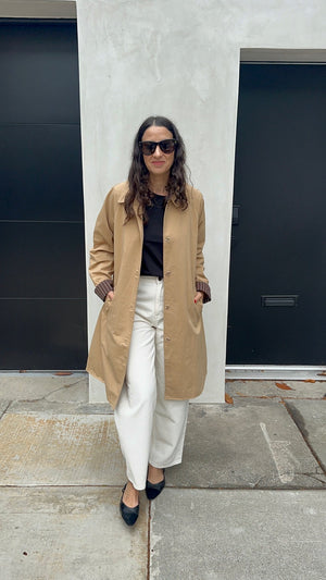 A woman stands outdoors in front of a concrete wall and black garage doors, wearing a long tan trench coat over a black top and white wide-leg pants with black pointed flats.