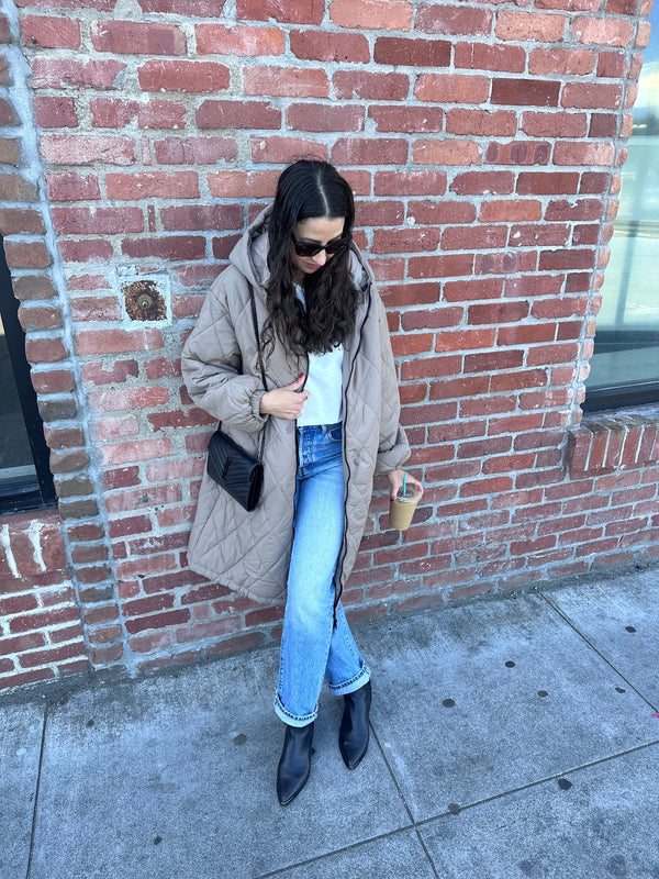 Woman in a beige coat standing against a brick wall, holding an ice cream cone.