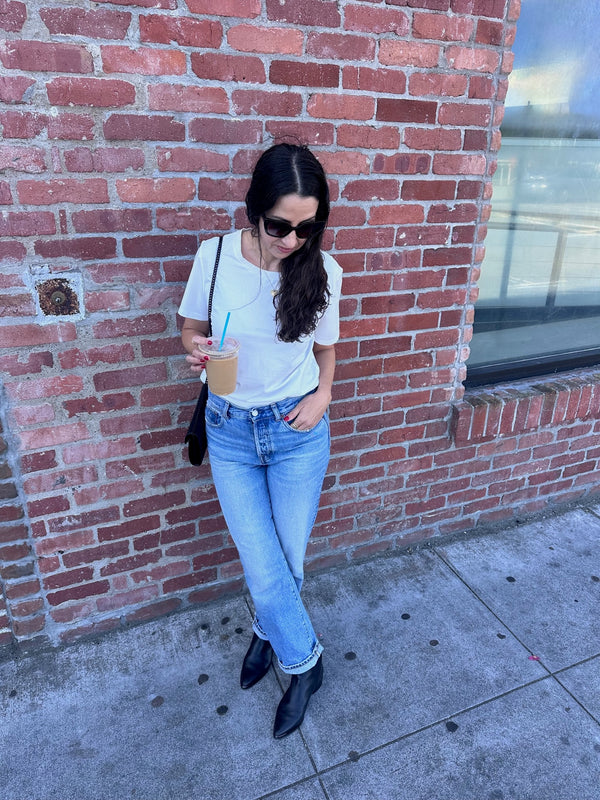 Woman standing against a brick wall holding a drink, wearing sunglasses and casual clothing.