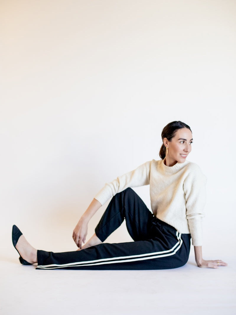 Woman in a white sweater and black pants with white stripes sitting on a white background