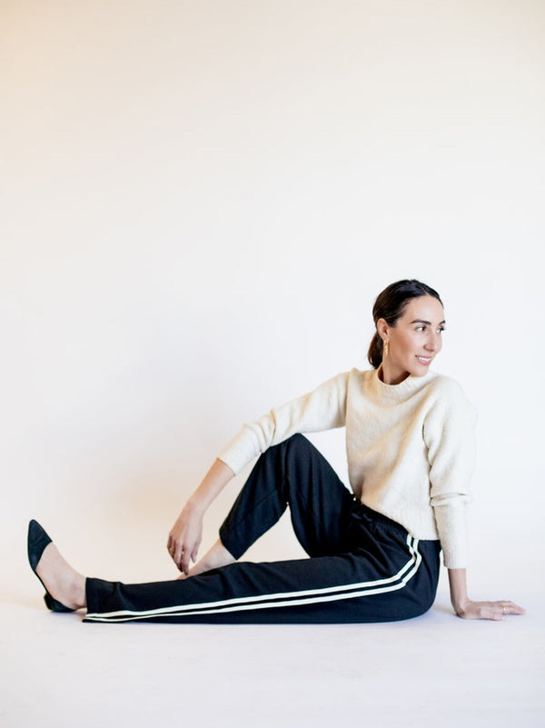 Woman in a white sweater and black pants with white stripes sitting on a white background