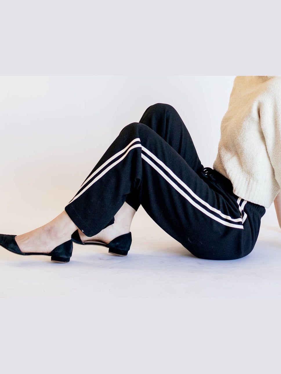 Person wearing black pants with white stripes sitting on a light-colored floor.