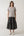 A woman stands against a plain light gray studio background wearing a light gray short-sleeve crewneck t-shirt and a dark charcoal gray tiered midi skirt, paired with white slide sandals.
