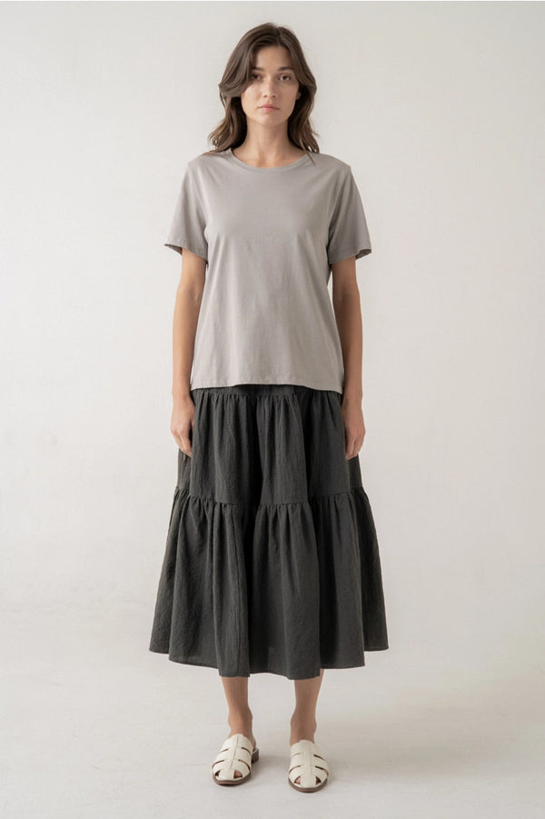 A woman stands against a plain light gray studio background wearing a light gray short-sleeve crewneck t-shirt and a dark charcoal gray tiered midi skirt, paired with white slide sandals.