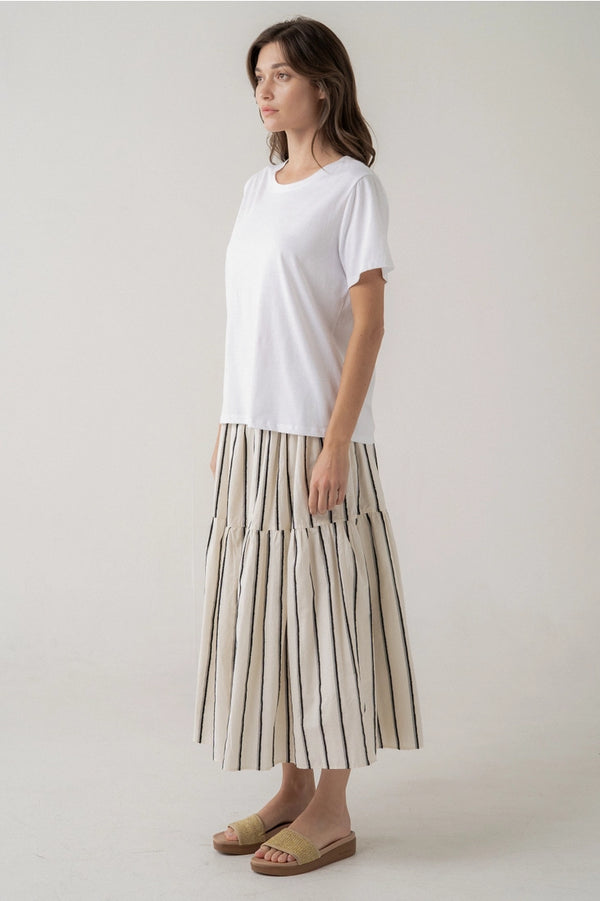 A woman modeling a white short-sleeve crewneck t-shirt and a tiered midi skirt with vertical beige and black stripes, paired with tan slide sandals, against a plain light gray studio background.