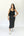 A woman modeling a black satin midi dress with a cowl neckline and thin straps, paired with white sneakers and a black shoulder bag, against a plain white studio background.