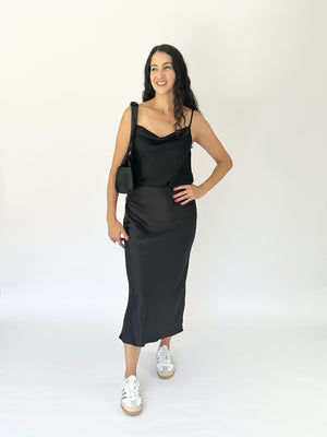 A woman modeling a black satin midi dress with a cowl neckline and thin straps, paired with white sneakers and a black shoulder bag, against a plain white studio background.