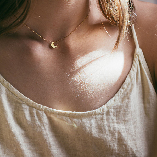 Close-up of a person wearing a gold necklace with a moon pendant, wearing a light-colored top.