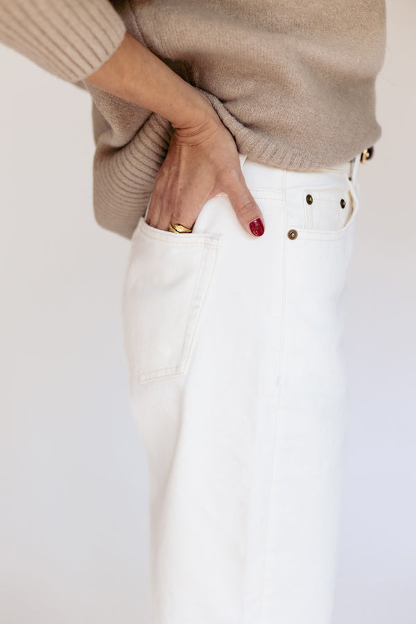 A close-up of a person wearing white high-waisted jeans with gold-tone rivets and a beige ribbed knit sweater, with one hand tucked into the back pocket.