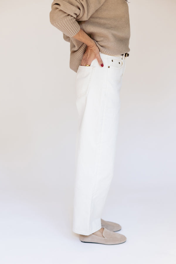 A person wearing white pants, a beige knit sweater, and beige slip-on shoes, standing against a plain light background.