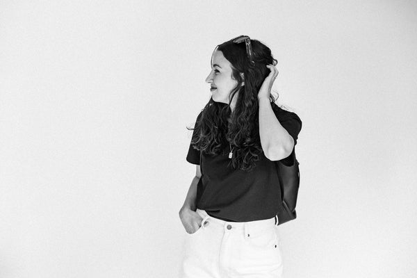 A woman with long dark hair stands in profile against a plain light background, wearing a black short-sleeve top and white pants, with sunglasses resting on her head.