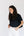 A woman with long, wavy dark brown hair wearing a black short-sleeve polo shirt and white pants, standing against a plain white studio background.