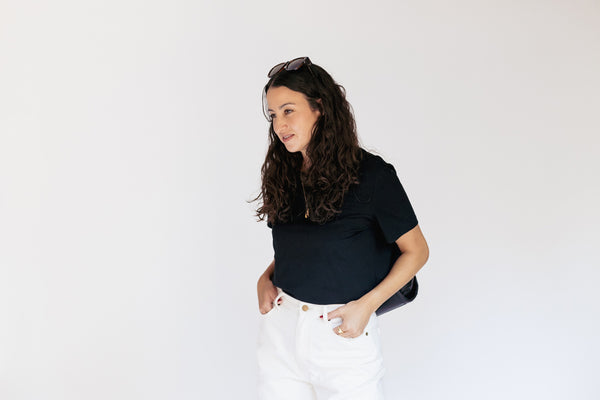 A woman with long, wavy dark brown hair wearing a black short-sleeve polo shirt and white pants, standing against a plain white studio background.