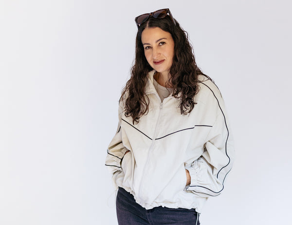 A woman with long, wavy brown hair wearing a cream-colored zip-up jacket with black piping details and dark jeans, standing against a plain white background.