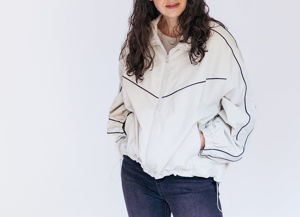 A woman wearing a cream-colored zip-up windbreaker jacket with dark blue piping details, paired with dark blue jeans, against a plain white background.