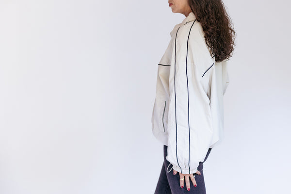 A woman shown from the side wearing a light beige or off-white hooded jacket with thin dark piping details, paired with dark pants, against a plain white studio background.