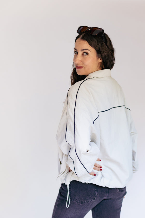 A woman wearing a white zip-up jacket with black piping details, paired with dark jeans, photographed against a plain white studio background.