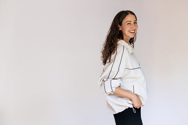 A woman with long dark hair is shown in profile against a plain light background, wearing a loose white shirt with thin dark piping and dark pants.
