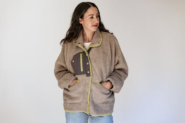 A woman wearing a taupe sherpa fleece jacket with a dark brown chest pocket and neon yellow piping, paired with blue jeans, against a plain white studio background.