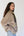 A woman wearing a beige sherpa fleece jacket with olive green trim and light blue jeans, standing against a plain white wall.