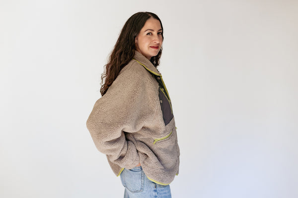 A woman wearing a beige sherpa fleece jacket with olive green trim and light blue jeans, standing against a plain white wall.