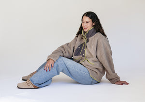 A woman sitting on a white studio floor wearing a beige sherpa fleece jacket with a dark gray collar and light olive green zipper accents, paired with light blue jeans and tan suede boots.
