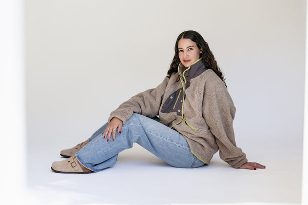 A woman sits on the floor in a studio setting, wearing a beige sherpa fleece zip-up jacket with dark gray panels and light green piping, paired with light blue jeans and tan mule shoes.