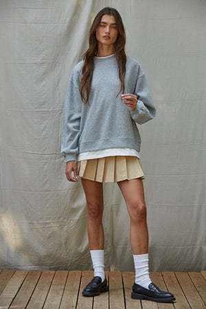 Woman wearing a gray sweater, beige pleated skirt, and white socks against a plain background