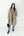 A woman modeling a long, quilted, taupe-brown hooded puffer coat over a brown and white striped knit sweater, blue jeans, and black ankle boots, holding a black handbag against a plain white studio background.