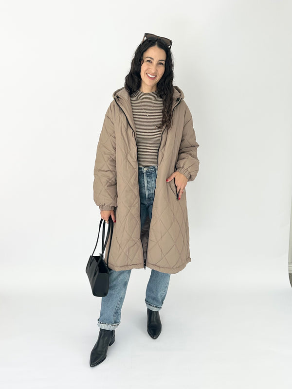 A woman modeling a long, quilted, taupe-brown hooded puffer coat over a brown and white striped knit sweater, blue jeans, and black ankle boots, holding a black handbag against a plain white studio background.