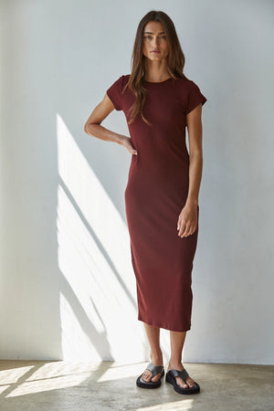 A woman modeling a short-sleeve, knee-length burgundy dress in a brightly lit room with natural window light casting shadows on the wall.