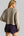 A woman shown from the back wearing a cropped, long-sleeve knit sweater in a light taupe-beige color and dark purple shorts.