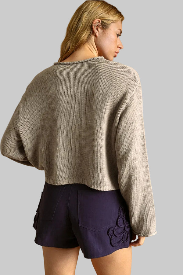 A woman shown from the back wearing a cropped, long-sleeve knit sweater in a light taupe-beige color and dark purple shorts.