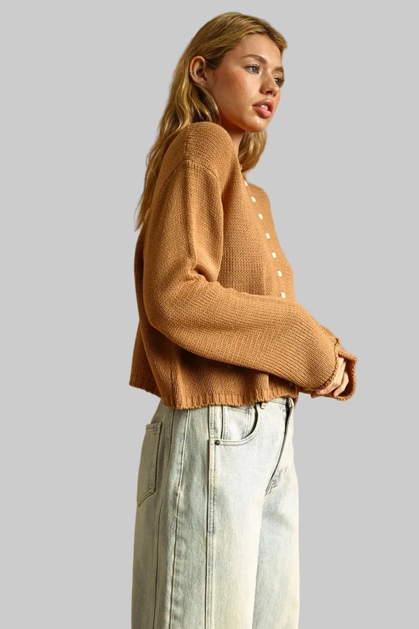 A woman wearing a cropped, long-sleeve knit cardigan in a warm camel brown color with white buttons, paired with light-wash wide-leg jeans.