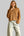A woman wearing a cropped, long-sleeve knit cardigan in a warm camel brown color with white buttons, paired with light-wash wide-leg jeans.