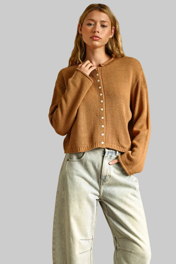 A woman wearing a cropped, long-sleeve knit cardigan in a warm camel brown color with white buttons, paired with light-wash wide-leg jeans.