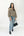A woman modeling a beige and black horizontally striped knit sweater, paired with light-wash blue jeans, black ankle boots, and holding a black handbag, against a plain white studio background.