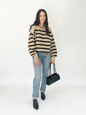 A woman modeling a beige and black horizontally striped knit sweater, paired with light-wash blue jeans, black ankle boots, and holding a black handbag, against a plain white studio background.