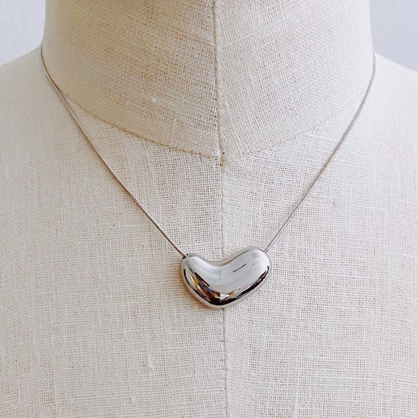 A polished silver heart-shaped pendant necklace displayed on a cream linen mannequin bust.