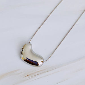 A polished metallic heart-shaped pendant necklace on a delicate silver chain, displayed on a light, textured white surface.