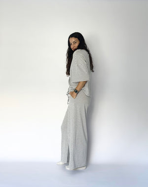 A woman with long dark hair stands in profile against a plain white studio background, wearing a loose white top and matching wide-leg pants with thin dark horizontal stripes.