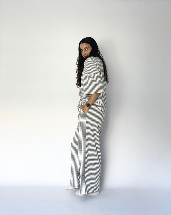 A woman with long dark hair stands in profile against a plain white studio background, wearing a loose white top and matching wide-leg pants with thin dark horizontal stripes.