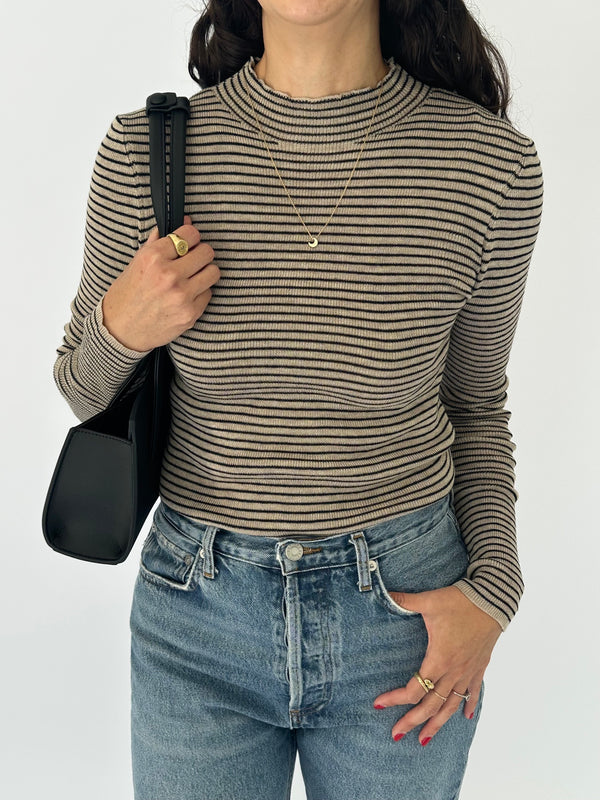 A woman wearing a black and taupe horizontally striped long-sleeve ribbed knit top, paired with blue jeans, a gold necklace, and a black shoulder bag, against a plain white background.