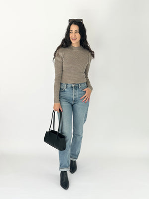 A woman modeling a black and taupe striped mock neck knit top, paired with light blue jeans, black ankle boots, and carrying a black handbag, set against a plain white studio background.