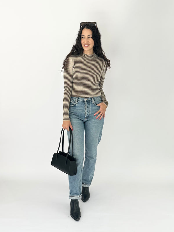 A woman modeling a black and taupe striped mock neck knit top, paired with light blue jeans, black ankle boots, and carrying a black handbag, set against a plain white studio background.
