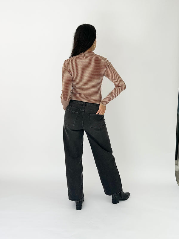 A woman is shown from behind wearing a red and beige horizontally striped long-sleeve turtleneck top, black wide-leg jeans, and black boots, standing against a plain white studio background.