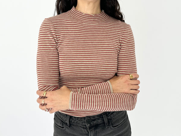 A person wearing a fitted long-sleeve mock neck top with thin horizontal red and off-white stripes, paired with dark gray jeans, against a plain white background.